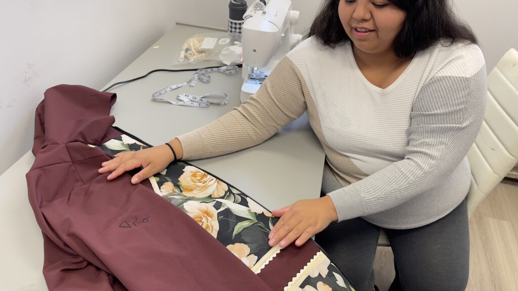 A woman with dark hair and a warm expression sits at a sewing workstation, carefully smoothing fabric on a maroon garment with floral and lace detailing. A sewing machine, measuring tape, and crafting supplies are visible on the table, indicating an ongoing design or tailoring project. The setting appears to be a creative workspace.