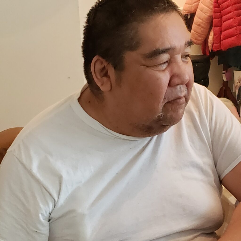 A middle-aged man with short dark hair and a slight smile sits indoors wearing a white t-shirt. He is looking off to the side with a calm expression. Behind him, jackets and bags hang on a rack. The lighting is soft and natural.