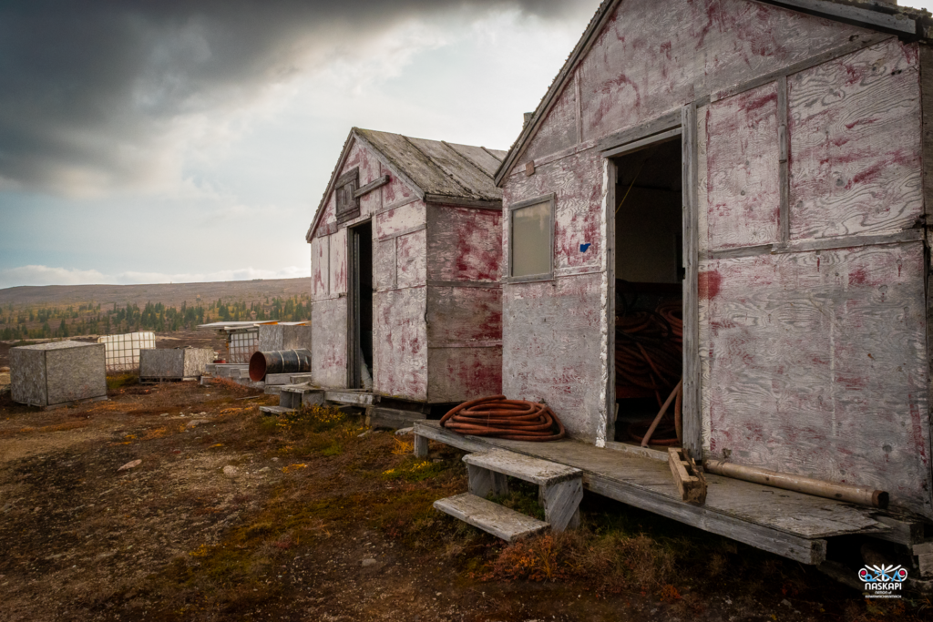 
The image depicts two weathered, wooden structures with peeling paint, showing a mix of gray and faded red hues. They appear to be makeshift buildings or storage sheds, with one door slightly ajar, revealing coiled hoses inside. The structures are elevated slightly off the ground on wooden blocks, with simple steps leading up to the doors. The surrounding area is a barren, rocky landscape with sparse vegetation, and several large, industrial containers and barrels are scattered nearby. In the distance, a line of evergreen trees stretches across the horizon under a cloudy sky, suggesting a remote, rugged location. The overall atmosphere is one of isolation, with the muted colors and worn materials conveying a sense of age and exposure to the elements. The image is marked with the Naskapi Development Corporation logo in the bottom right corner.