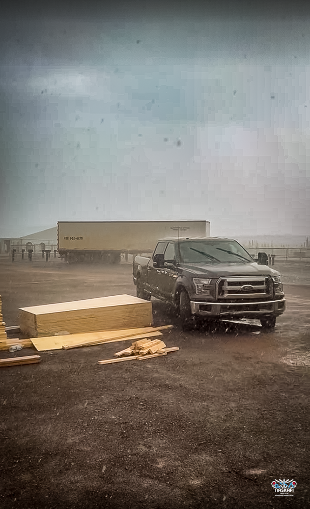 The image shows a construction site on a rainy day, with a gray Ford pickup truck parked on wet dirt. The sky is overcast with heavy clouds, and rain is visibly falling, creating a misty atmosphere. In front of the truck, several large wooden boards and construction materials are scattered on the ground, some partially covered by rain. In the background, a large beige shipping container trailer is parked, with its side partially obscured by the rain. The area appears to be a staging ground for construction, with materials and equipment arranged around the site. The ground is muddy and wet, reflecting the dreary weather. The image is marked with the Naskapi Development Corporation logo in the bottom right corner.