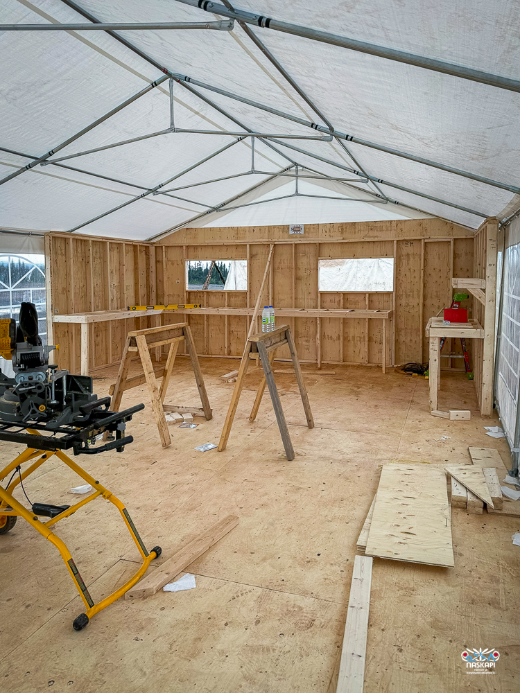 The image shows the interior of a temporary construction shelter, likely serving as a workspace or kitchen under construction. The structure is framed with wooden walls, and the roof is covered by a white tarp supported by metal rods. The floor is made of plywood, with various construction materials and tools scattered around. In the center of the space, two wooden sawhorses stand, with a level tool resting on one of them. To the left, there is a miter saw mounted on a yellow stand, ready for use. The walls have small rectangular windows cut out, allowing some natural light to enter. The overall space appears organized but unfinished, with tools and materials suggesting ongoing work. The image is marked with the Naskapi Development Corporation logo in the bottom right corner.