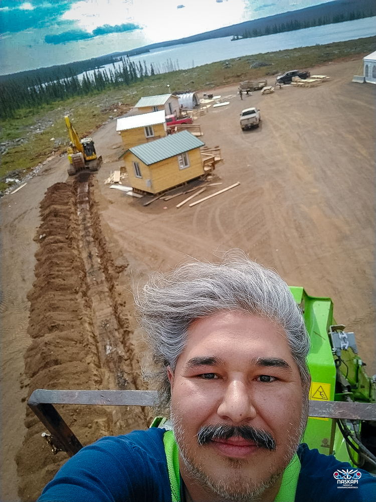 The image features a selfie of a person with gray hair and a mustache, taken from an elevated position, possibly on a construction vehicle. The person is smiling slightly, with a backdrop of a construction site below. The site includes several small wooden buildings with green metal roofs, scattered across a large, dirt-covered area. In the background, a yellow excavator is seen digging a long trench, with piles of earth on either side. Further back, a large, serene lake stretches across the horizon, surrounded by dense evergreen trees. The sky above is partly cloudy, with patches of blue visible. The overall scene suggests ongoing construction work in a remote, picturesque location. The image is marked with the Naskapi Development Corporation logo in the bottom right corner.