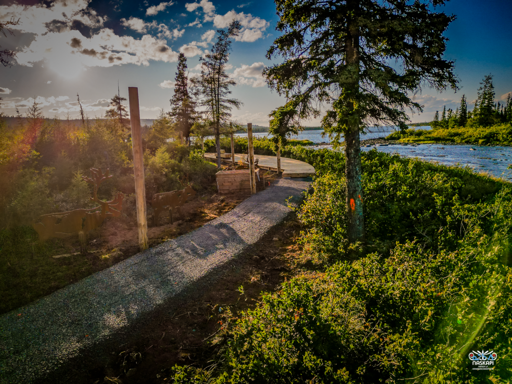 A path under construction, with wooden beams laid along a gravel walkway. Caribou sculptures stand to the side of the path, while the river flows in the background. The trees and bushes surrounding the area are bathed in the warm light of the setting sun.