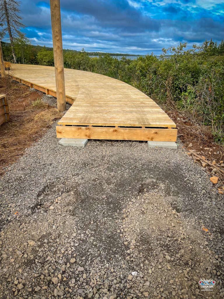 A newly constructed section of the boardwalk, curving gently to the left, surrounded by lush greenery. The wooden planks are freshly installed, but the gravel path leading up to the boardwalk shows signs of disturbance. The sky is overcast, with dense clouds looming overhead. This area is part of an ongoing construction project.