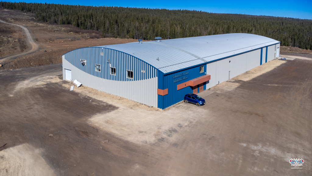 The image shows a large, blue and gray metal building labeled "Shinasheshtooan Memorial Arena" on the side. A single blue vehicle is parked near the front entrance, which has orange accents. The building sits in an open area with a dirt parking lot, surrounded by rugged terrain and a dense forest of evergreen trees in the background. The overall setting suggests a remote location. The logo of the Naskapi Nation of Kawawachikamach is visible in the lower right corner.