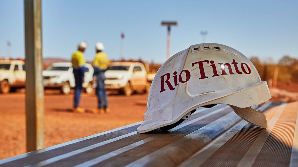 This image shows a close-up of a white hard hat with the "Rio Tinto" logo in bold red letters resting on a metallic surface. In the background, two workers in high-visibility clothing and hard hats are seen standing near several white utility vehicles on a dusty, outdoor worksite, likely a mining location. The workers appear to be discussing something, with the vehicles and red earth suggesting a remote or industrial setting related to mining or resource extraction. The hard hat in the foreground emphasizes safety and the company's branding.