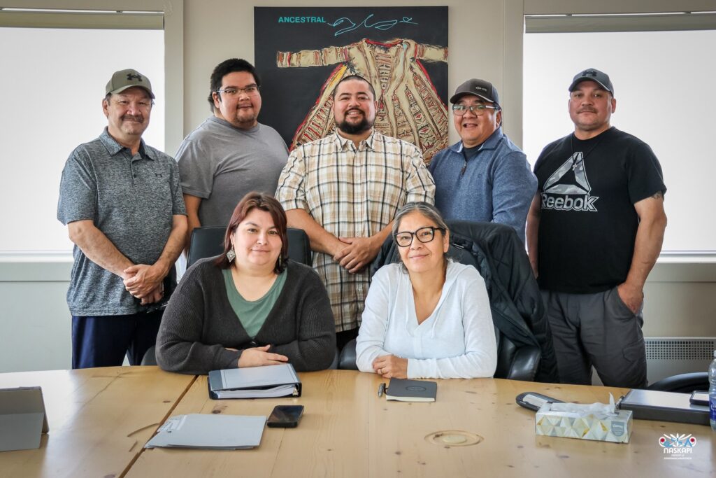 This image shows a group of seven people, five men standing and two women seated at a table, posing for a group photo. They are indoors, with a framed artwork of a traditional garment labeled "Ancestral" hanging on the wall behind them. The individuals are smiling, and some are dressed casually, with one person wearing a cap and another in a Reebok T-shirt. On the table in front of them are notebooks, a tablet, and a phone. The logo in the bottom right corner features the word "Naskapi."