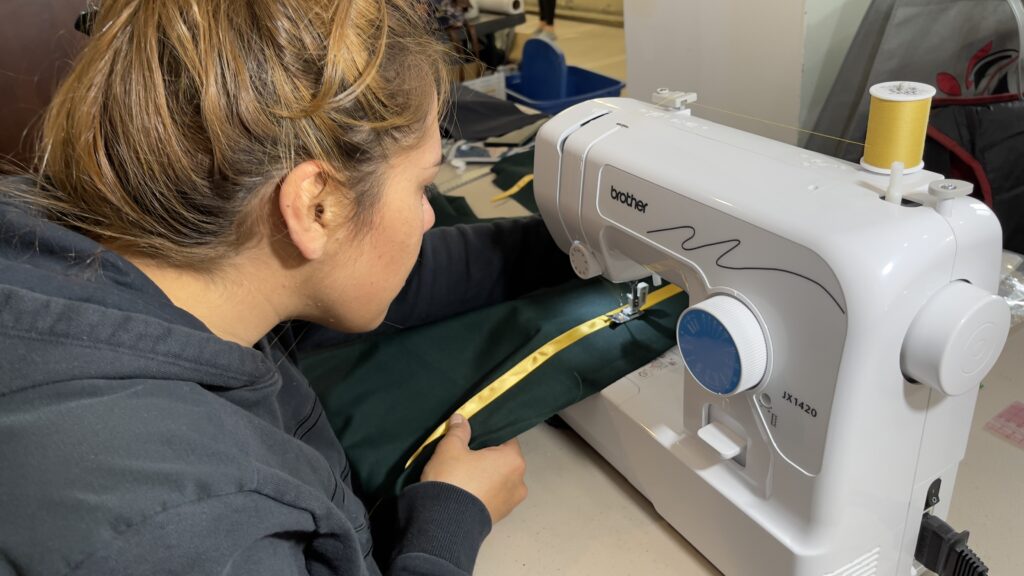 A young woman with light brown hair tied back is focused on sewing a green fabric with a gold ribbon trim using a Brother JX1420 sewing machine. She wears a black hoodie and leans in closely as she guides the fabric under the needle. The workspace around her contains various sewing materials, including fabric pieces, a spool of yellow thread, and tools for garment construction.